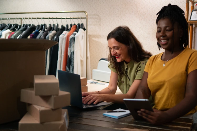 Two women smiling and preparing clothing for packaging in an e-commerce thrift store