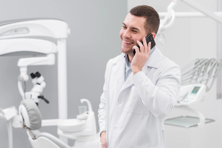 Smiling male dentist in a white coat talking on a smartphone in his dental clinic.
