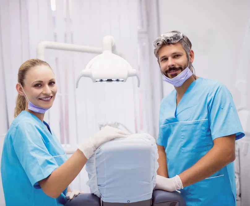 Two smiling dentists in light blue scrubs and masks standing next to a dental chair in a bright, modern clinic.